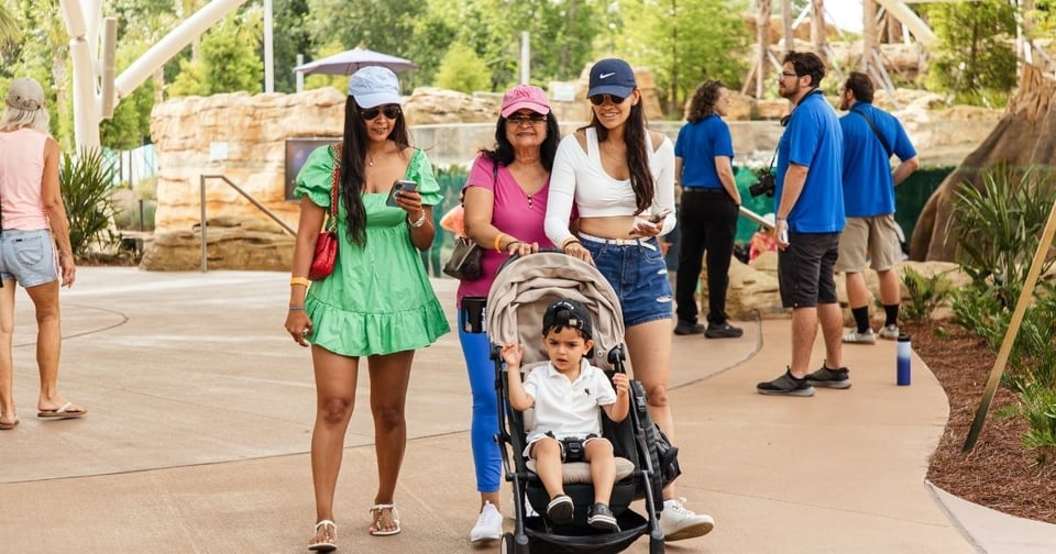 Three women walking through a zoo with a young child in a stroller, while staff members stand near an animal habitat in the background.